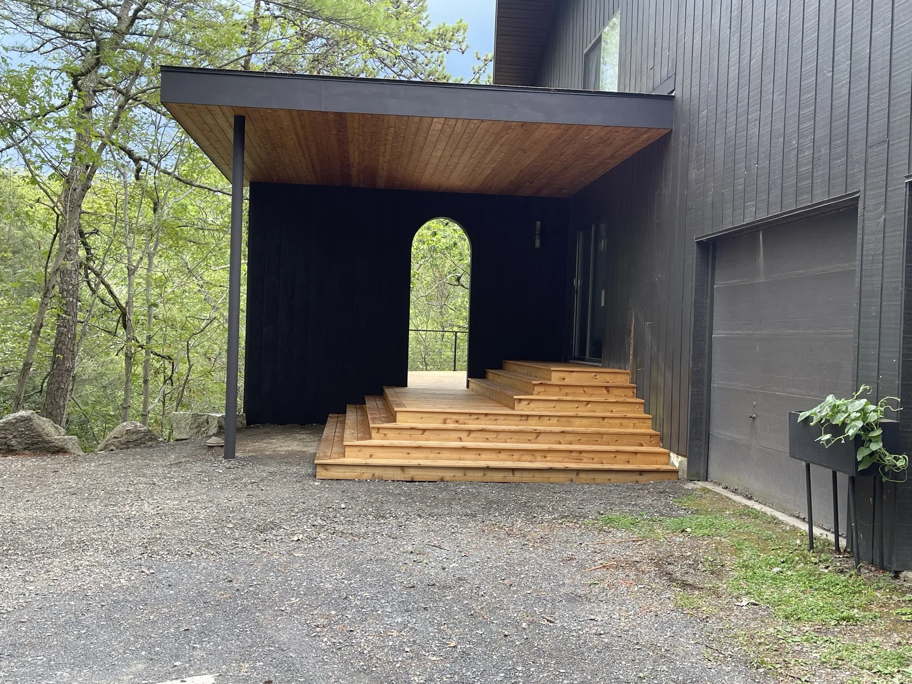 Wide cedar entry stair and landing beneath a cedar-clad soffit on a dark board-and-batten house, with an arched pass-through beyond