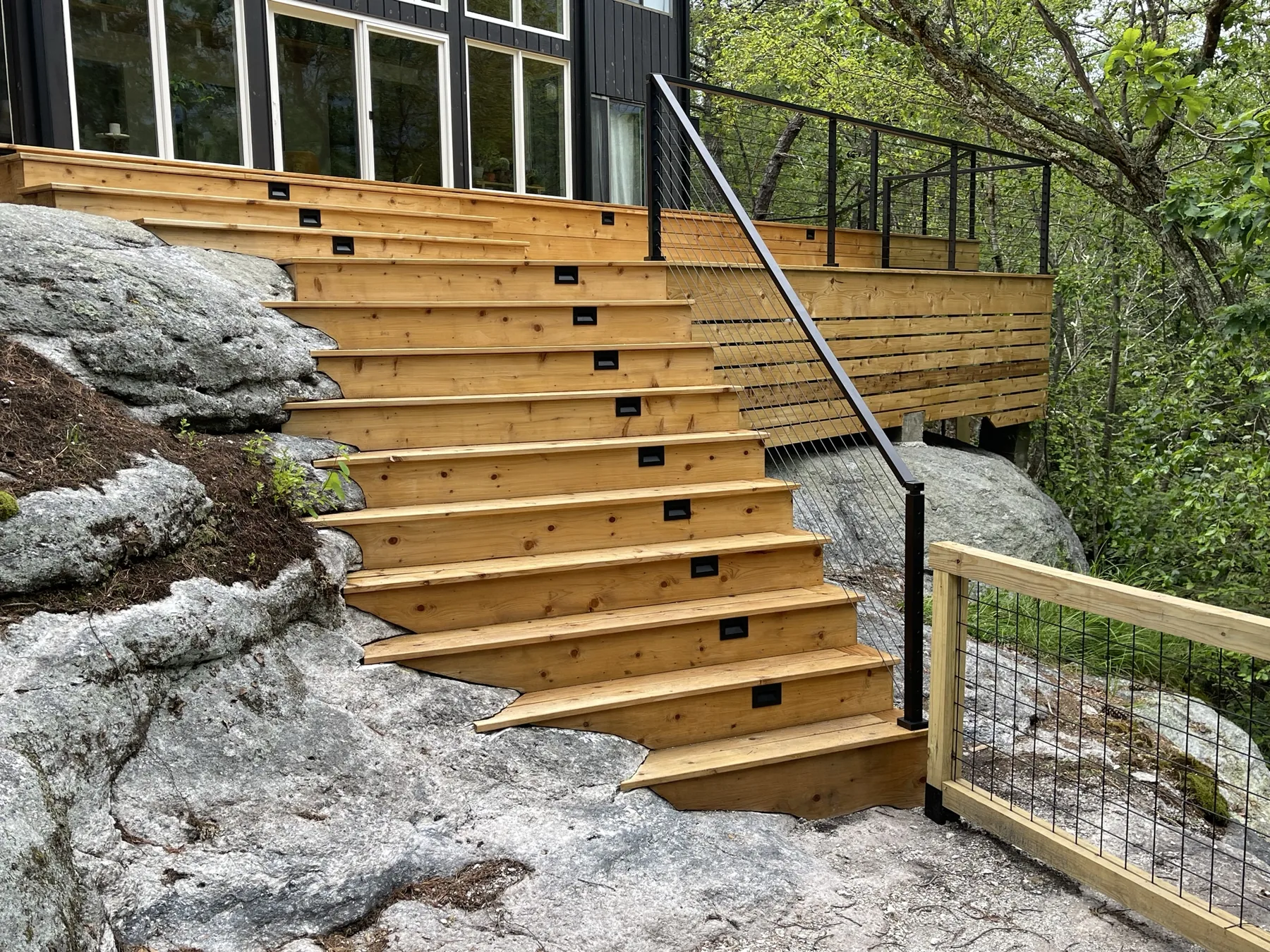 Wide cedar stair scribed to the natural bedrock, with integrated riser lights and a black cable railing, leading up to a deck at a dark-sided house