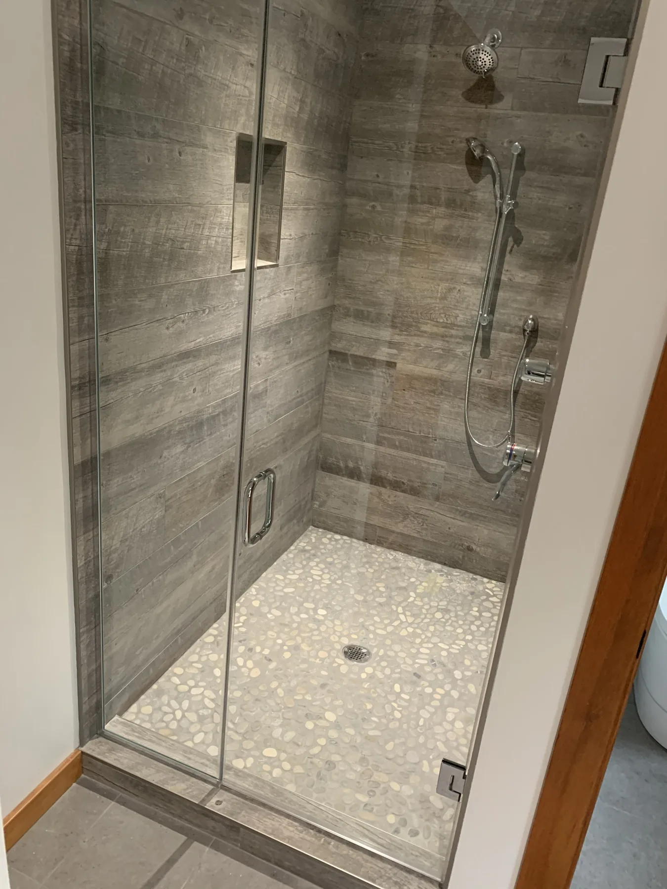 Glass-door walk-in shower tiled in wood-look grey porcelain with a pebble floor and recessed niche